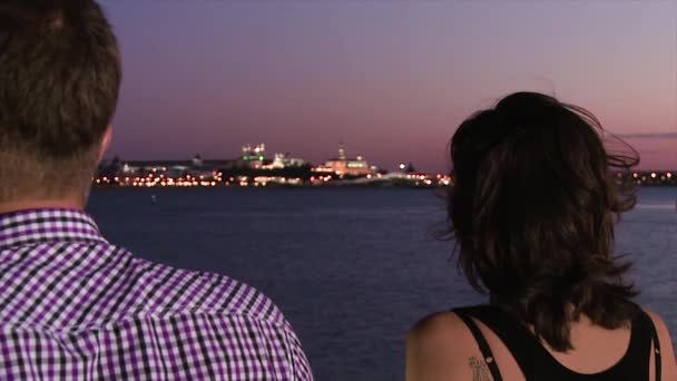 站在码头上的男人和女人。现场。从站在码头上的一男一女的背后特写镜头。从码头到另一边的闪亮城市的夜景与日落色的天空 — 图库视频影像
