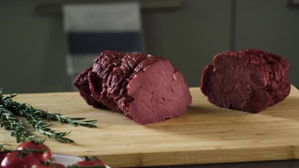 Close-up de cortar pedaço de presunto. Cena. Corte por mãos de mulheres em duas partes pedaço de presunto suculento. Presunto fatiado em tábua de madeira no fundo de ramos verdes de verdes e tomates — Vídeo de Stock