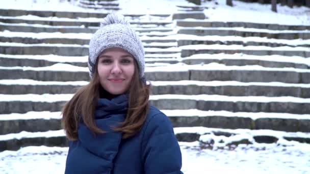 Mulher bonita no chapéu de inverno sorri de pé do lado de fora na neve na floresta com fundo de escadas nevadas. Retrato de uma menina bonita olhando para a câmera. Retrato de inverno de uma jovem. O — Vídeo de Stock