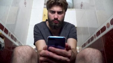 Bir adam tuvalette otururken zaman geçirmek için onun smartphone tutar. Klozet üzerinde evde otururken cep telefonu kullanan genç adam. Cep telefonu oynarken klozet üzerinde oturan adam