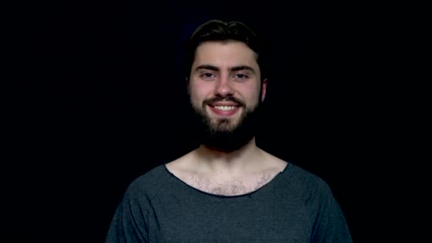 Foto de cerca de un hombre barbudo sonriente feliz, aislado sobre un fondo negro. Hombre guapo con camiseta gris y sonriente — Vídeo de stock