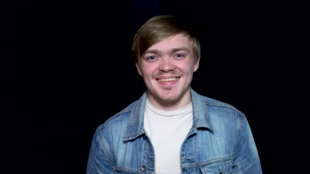 Jonge knappe model op zwarte achtergrond in een jean jas geeft een vrolijke glimlach. Jonge, bebaarde man geeft een grote glimlach. — Stockvideo