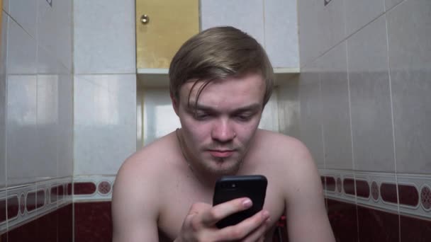 若い男は、我が家のトイレの上に座って携帯電話を使用します。携帯電話をトイレに坐っていた男。スマート フォンへの依存 — ストック動画