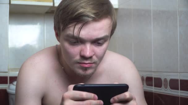Молодой человек пользуется мобильным телефоном, сидя дома на унитазе. Мужчина сидит в туалете с телефоном. Крупный план человека с щетиной, играющего в телефоне на унитазе. Зависимость от смартфонов — стоковое видео