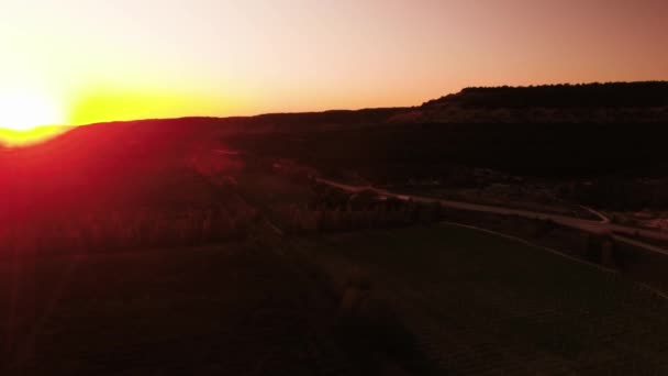 赤い夕日。ショット。山によって部分的に隠された太陽の明るいディスクと赤い夕焼け空。日没の農村フィールドを通る地方のハイウェイにトラフィックのトップ ビュー — ストック動画