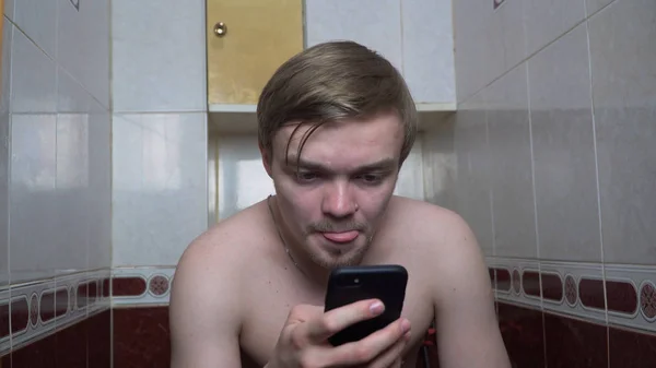 Le jeune homme utilise un téléphone portable assis sur les toilettes à la maison. Homme assis dans les toilettes avec téléphone. Dépendance sur les smartphones — Photo