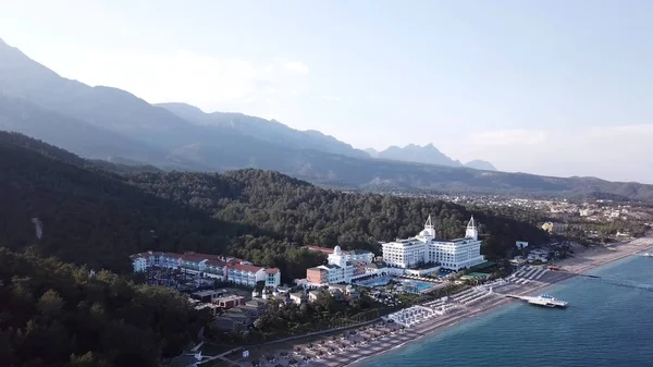 Video. Aerial view of Hotel on sea shore and mountains covered with green forest, trees with blue sky at sunrise. Video. Aerial view on seashore with luxury Hotel with forest and mountains background