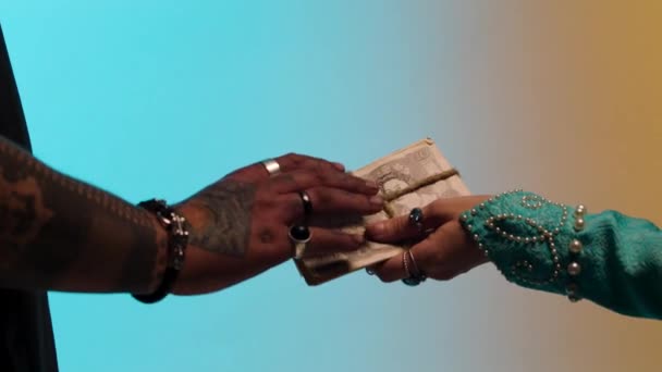 Hands of eastern woman with many rings giving money tied up with twine to seller man, isolated on yellow and blue background. Stock. Woman paying by cash to a seller man, purchasing concept. — Stock Video