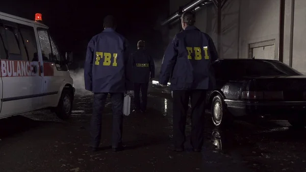 FBI agenti pracují na místě činu v noci, policejní auto s světla a sanitka pozadím. Zadní pohled na tři agenti Fbi jdou na kriminální scény — Stock fotografie