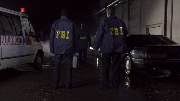 Агенты ФБР работают на месте преступления ночью, полицейская машина со светом и биографией скорой помощи. Взгляд сзади на трех агентов ФБР идет к месту преступления

