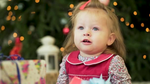 Mooi klein meisje in de buurt van het nieuwe jaar boom. Meisje glimlacht en spelen met giften op de achtergrond van kerstbomen. Mooi meisje in jurk in de buurt van de kerstboom. — Stockvideo