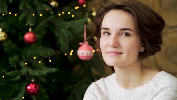 Concepto de Navidad, hermosa morena sonriendo en el fondo de un árbol de Navidad decorado creativamente. Retrato de cerca de la hermosa joven sonriendo a la cámara con el árbol de Navidad detrás — Vídeos de Stock