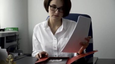 Beyaz ofis duvar arka plan üzerinde kırmızı klasör belgelerden düzenleme bardaklarda kısa, koyu renk saçlı güzel kadın. Beyaz gömlekli kağıtları, iş kavramı ile çalışan hanım konsantre.