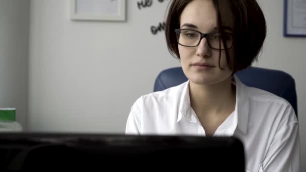 Bliska portret pięknej młodej kobiety w białej koszuli, patrząc na ekran laptopa na tle ściany biały biuro. Nowoczesna kobieta interesu pracę przed ekranem komputera, w jej miejscu pracy. — Wideo stockowe