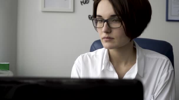 Närbild porträtt av en vacker ung kvinna i vit skjorta tittar på laptop skärmen på vita kontor vägg bakgrund. Modern affärskvinna arbetar framför datorskärmen i hennes arbetsplats. — Stockvideo