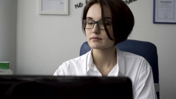 Primer plano para la mujer joven en camisa blanca mirando a la computadora portátil, contestando el teléfono, y sonriendo en el fondo de la pared de la oficina blanca. Empresaria que trabaja delante de la pantalla del ordenador en su lugar de trabajo . — Vídeos de Stock