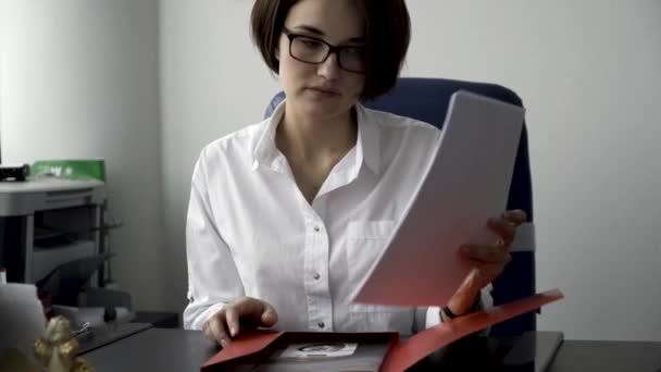 Vacker kvinna med kort, mörkt hår i glasögon att ordna dokument från röd mapp på vita kontor vägg bakgrund. Koncentrerad office lady i vit skjorta arbetar med papper, affärsidé. — Stockvideo