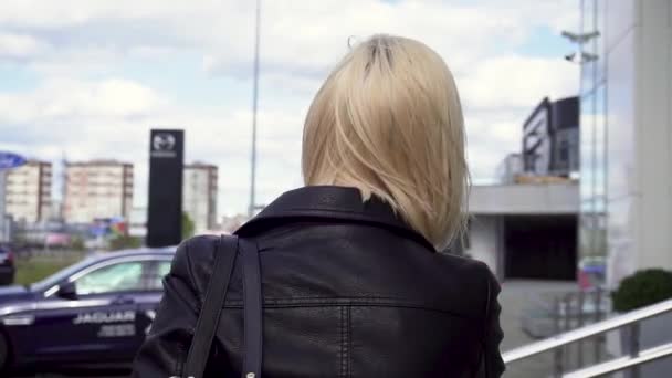 Rückansicht einer Blondine in schwarzer Lederjacke, die auf ein Gebäude mit Glasfassade zugeht. Aktien. Nahaufnahme für junge, blonde Frau mit einer Tasche über der Schulter, die ihre Haare berührt. — Stockvideo