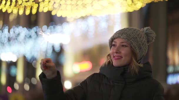 Retrato de una modelo de chica se ve con chispas en sus manos. Joven mujer sostiene chispeante en la noche, luces de Navidad en el fondo — Vídeos de Stock