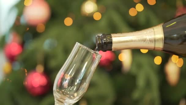 Detail šampaňské nalil do sklenice, vánoční strom na pozadí. Detail vylévání šampaňského na sklo vedle vánoční stromeček — Stock video