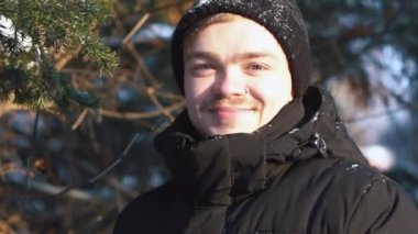 Gülümseyen genç adam kış ormanda kalmak ve kameraya seyir siyah ceket Close-up. Güneşli kış günü.