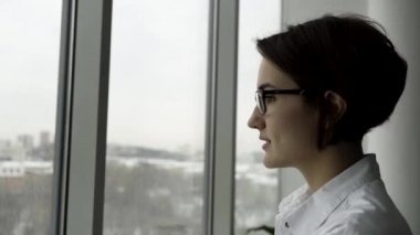 Zarif bir kadın ofis pencere tarafından ayakta ve gözlüklerini bulanık gri şehir manzaralı ayarlama kısa, siyah saçlı ile yan görünüm. Beyaz gömlek gözlüklerini dokunmadan iç geçiriyorum, yorgun hanım