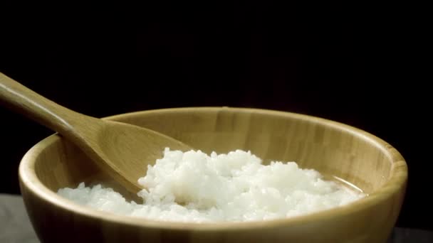 Cierre para el arroz al vapor en un tazón de madera con cuchara de madera, aislado sobre fondo negro. Arroz recién cocido con vapor en plato marrón profundo con cuchara grande, comida, concepto de cocina . — Vídeo de stock