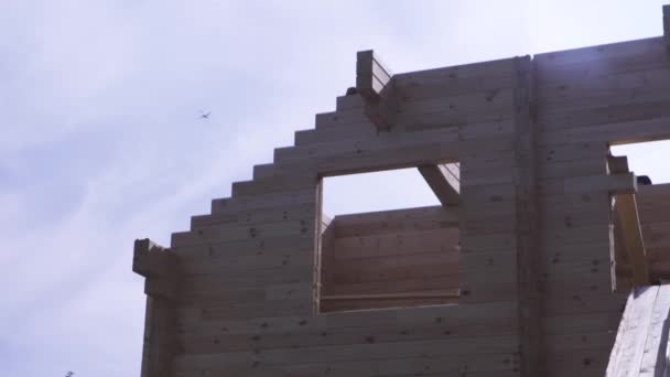 Frame of building, wooden log house on blue sky background with a plane flying away. Construction in progress of a wooden house against cloudy sky. — Stock Video