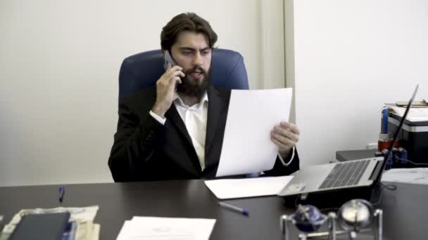 Концентрований, нервовий, молодий, бородатий бізнесмен по телефону, сидячи в синьому шкіряному кріслі в офісі. Злий чоловік у костюмі розмовляє по телефону і дивиться через документи . — стокове відео