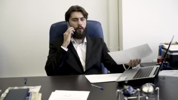 Empresario concentrado, nervioso, joven, barbudo por teléfono, sentado en sillón de cuero azul en la oficina. Hombre enojado en traje hablando por teléfono y mirando a través de documentos . — Vídeos de Stock