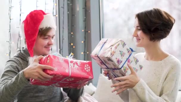 Feche para o casal trocando presentes enquanto celebram o Natal e o Ano Novo juntos. Homem em chapéu de Papai Noel e mulher dando presentes em papel de embrulho colorido e abraço, conceito de férias de inverno . — Vídeo de Stock