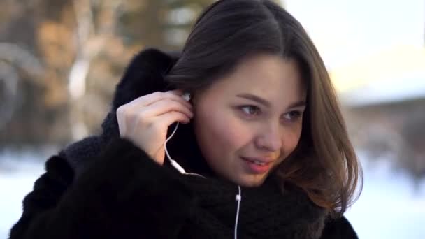 Mulher elegante, jovem com cabelo curto e escuro colocando fones de ouvido brancos enquanto caminha no parque de inverno. Menina bonita e atraente usando fones de ouvido enquanto caminhava ao ar livre no inverno . — Vídeo de Stock