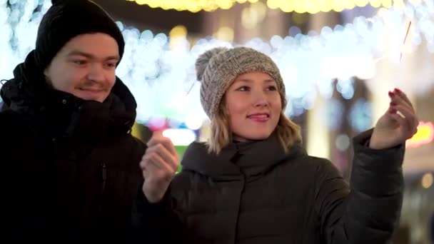 Coppia amorevole con scintille sorridenti, guardando negli occhi in città d'inverno di notte, Buon Natale e concetto di Capodanno. Uomo e donna con fuochi del bengala fanno contatto visivo su sfondo luci sfocate . — Video Stock