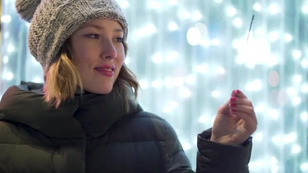 Close-up voor glimlachen, mooie meisje in de straat in de winternacht met sparkler over vervagen, blauwe lichten achtergrond. Vrolijke, mooie vrouw vasthouden van Bengalen brand en Nieuwjaar vieren. — Stockvideo