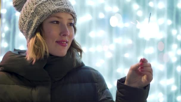 De cerca para sonreír, chica bonita en la calle en la noche de invierno con chispa sobre borroso, fondo de luces azules. Mujer alegre, hermosa sosteniendo fuego de bengala, celebrando el Año Nuevo . — Vídeos de Stock