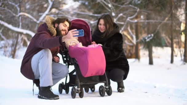 Joyeux, jeune famille marchant, faisant des photos dans un parc d'hiver, maman, papa et bébé en poussette. L'homme fait selfie wirh sa femme souriante et bébé en poussette rose sur fond d'arbres enneigés . — Video
