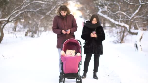 母と父は、冬の公園を歩きながら、ベビーカーで赤ちゃんを無視して携帯電話にはまって。親のベビーカー、中毒、値の概念で、幼い子供を無視してスマート フォンで忙しい. — ストック動画