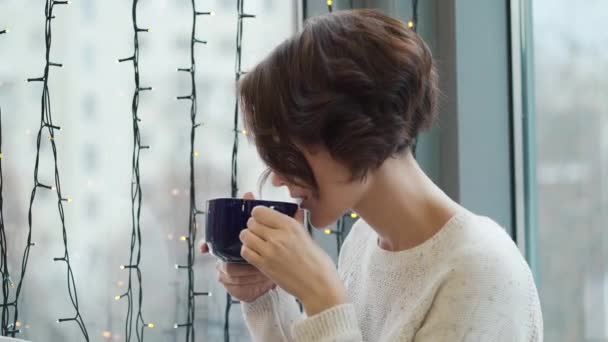 Zbliżenie: brunetka dziewczynka z filiżanką kawy w pobliżu okna, w przytulnej sali. Świąteczny nastrój. — Wideo stockowe