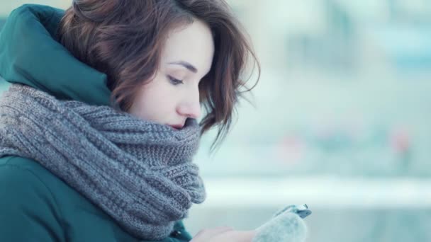 Close-up van jonge brunette meisje met een mobiele telefoon, zittend op een bankje in de winter. Centrum van de stad. — Stockvideo