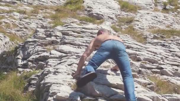 Weergave van de mens van rug stenen heuvel klimmen. Voorraad. Man in spijkerbroek en shirtless klimt stenen heuvel hellingen met groen gras — Stockvideo