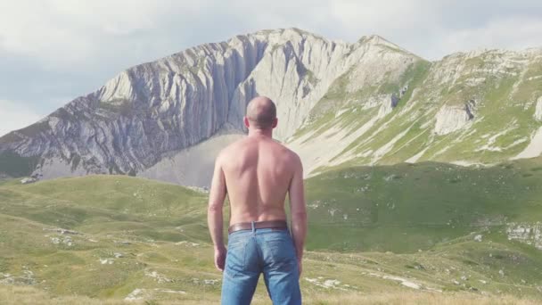 Vista da parte de trás do homem olhando para as montanhas. Estoque. Homem de jeans e sem camisa, encantado, olha para a rocha. Imagem corajosa e heróica do homem no fundo da paisagem poderosa de montanhas — Vídeo de Stock