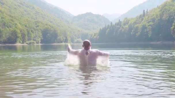 Чоловік у сорочці вистрибує з бризок води. Запас. Чоловік у білій сорочці плаває в гірському озері. Літні канікули в горах біля озера — стокове відео