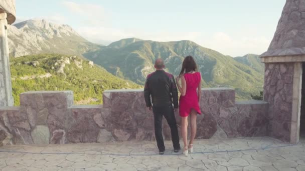 Πίσω όψη του ζευγάρι ψάχνει από το φρούριο βουνό. Απόθεμα. Ημερομηνία του ανθρώπου σε δερμάτινο σακάκι με σέξι γυναίκας με κόκκινο φόρεμα ενάντια στα βουνά. Εραστές παρατηρώντας ρομαντική θέα — Αρχείο Βίντεο