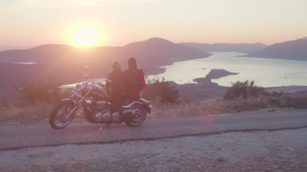 Vy från baksidan av romantiska par med motorcykel i solnedgången. Lager. Romantisk datum motorcyklist och kvinna i röd klänning på bakgrund av solnedgången och berg landskap med flod — Stockvideo