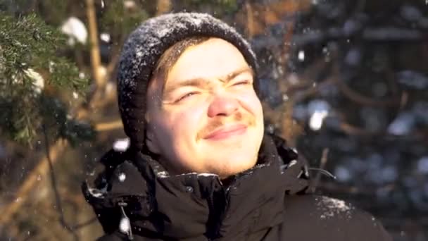 Retrato de un hombre joven y sonriente con bigote disfrutando de las nevadas en el bosque invernal, entrecerrando los ojos del sol brillante. Guapo, feliz hombre le gusta caer nieve sobre su cabeza en invierno día soleado . — Vídeo de stock