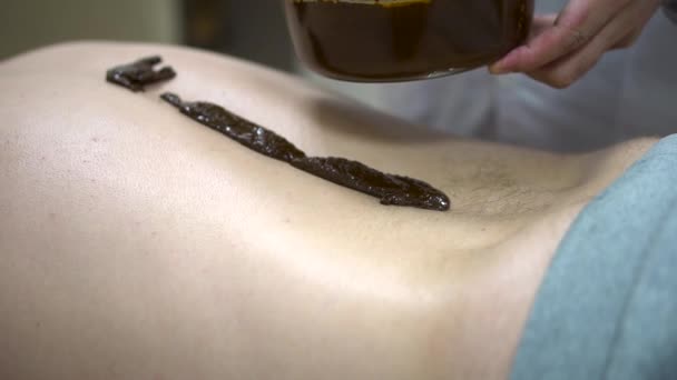 stock video Close up for a patient lying on a bed with hot therapeutic mud on his back, and alternative medicine concept. Close up for male back with hot, liquid herbs mixture, wellness procedure.