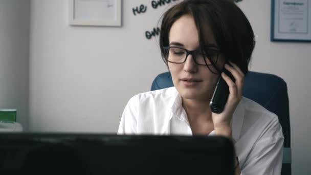 Segretario chiama al telefono e sorride in ufficio contro il suo computer portatile. Primo piano . — Video Stock