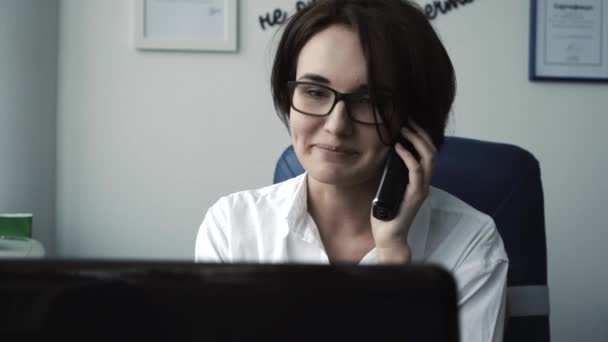 Secretaria llamando por teléfono y sonriendo en la oficina contra su computadora portátil. Primer plano . — Vídeos de Stock