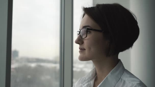 Affärskvinna i vit skjorta står nära fönstret och tittar på kameran i office. Närbild. — Stockvideo