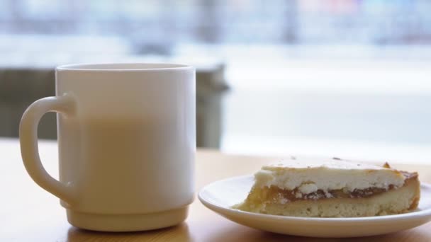 Close-up of white mug and plate with cheesecake. Morning breakfast with coffee and classic cheesecake on wooden table. Sweet breakfast in cafe on background of window with busy street — Stock Video